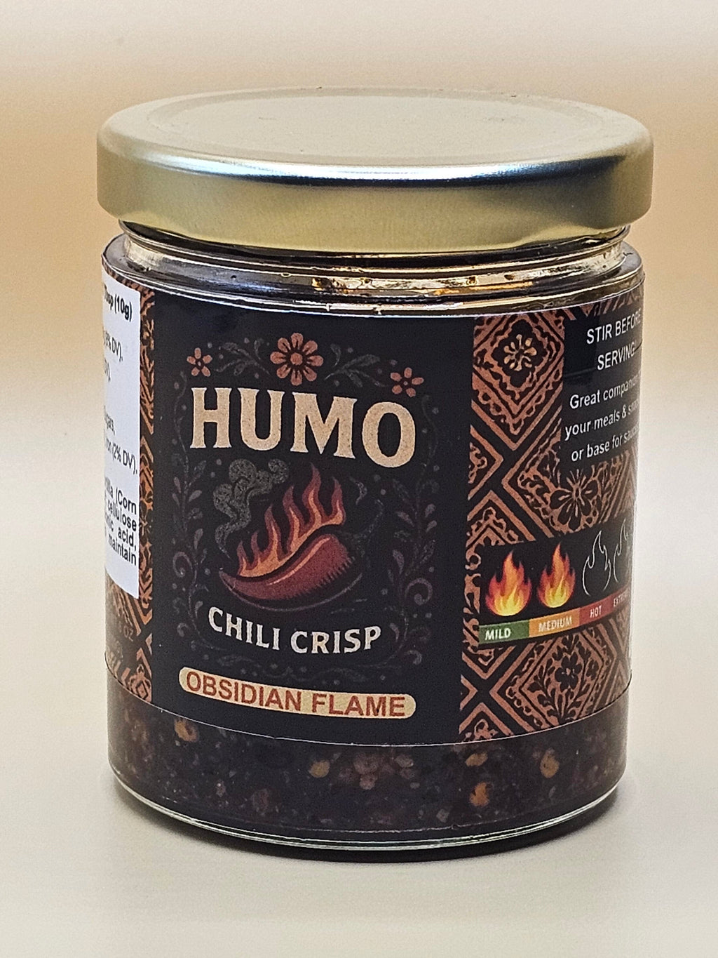 A glass jar of Humo Chili Crisp seasoning by Ossidian Spices, featuring a dark brown label with traditional patterns and a silver lid, photographed on a plain off-white background.