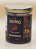 A glass jar of Humo Chili Crisp seasoning by Ossidian Spices, featuring a dark brown label with traditional patterns and a silver lid, photographed on a plain off-white background.