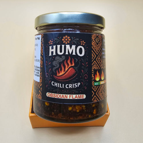 A glass jar of HUMO Chili Crisp Guelag Flame seasoning, featuring a dark label with orange and white text, resting on a light beige surface.