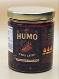 A jar of HUMO Chili Crisp Osidán Inferno hot sauce, featuring a dark brown label with gold lettering and a silver metal lid, set against a plain off-white background.