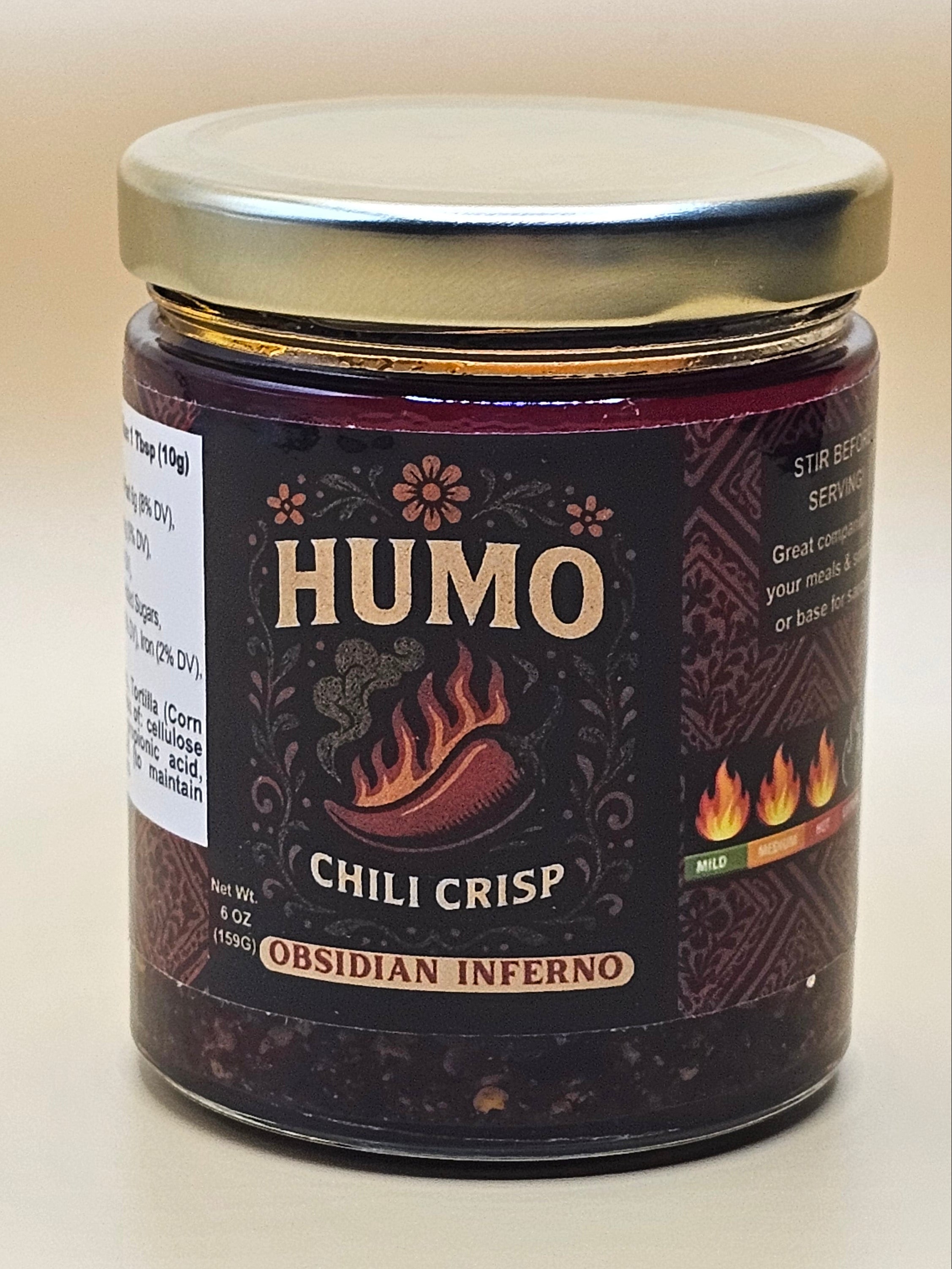 A jar of HUMO Chili Crisp Osidán Inferno hot sauce, featuring a dark brown label with gold lettering and a silver metal lid, set against a plain off-white background.