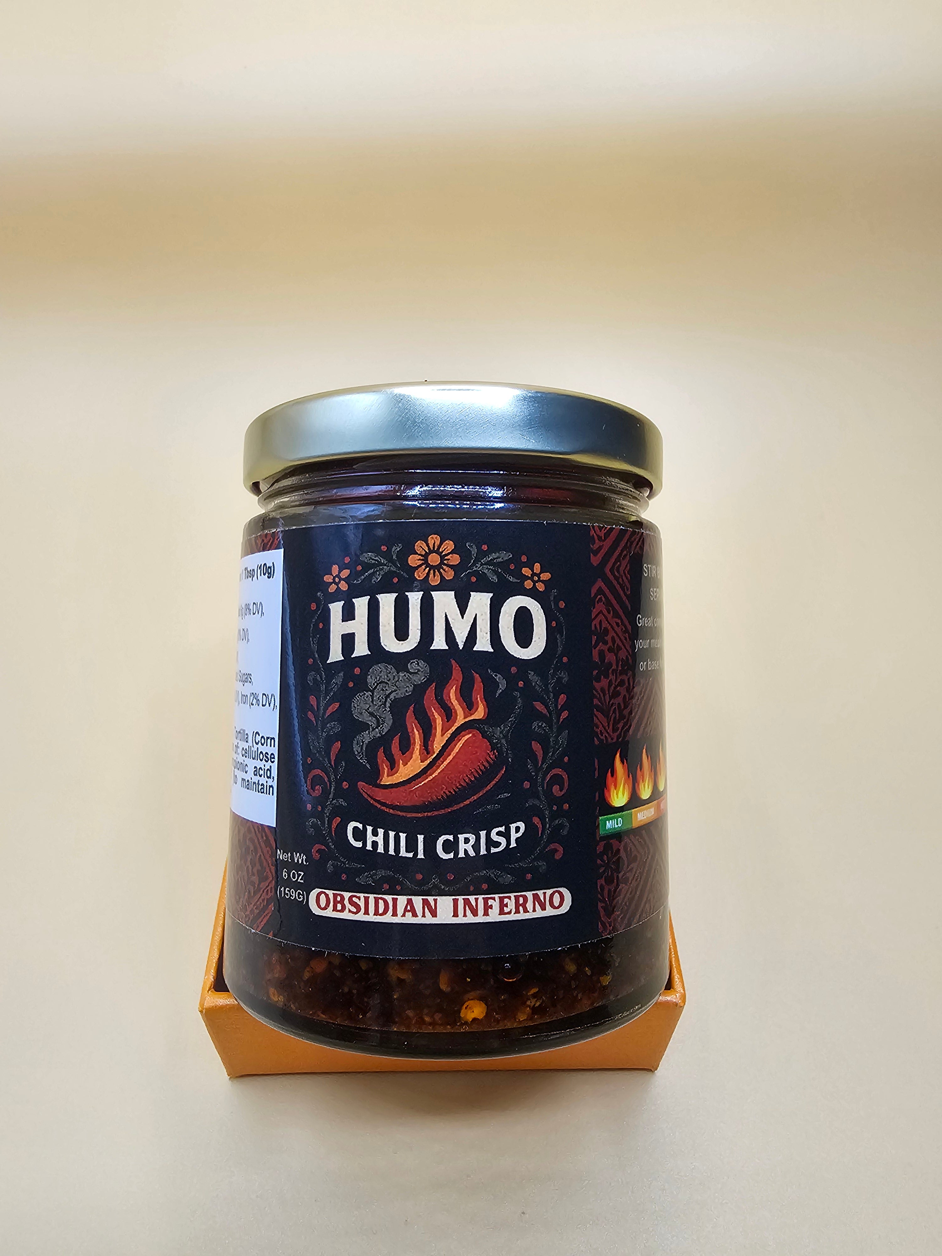 A glass jar of HUMO Chili Crisp Ossidian Inferno hot sauce, displayed on a small wooden shelf against a plain beige wall.